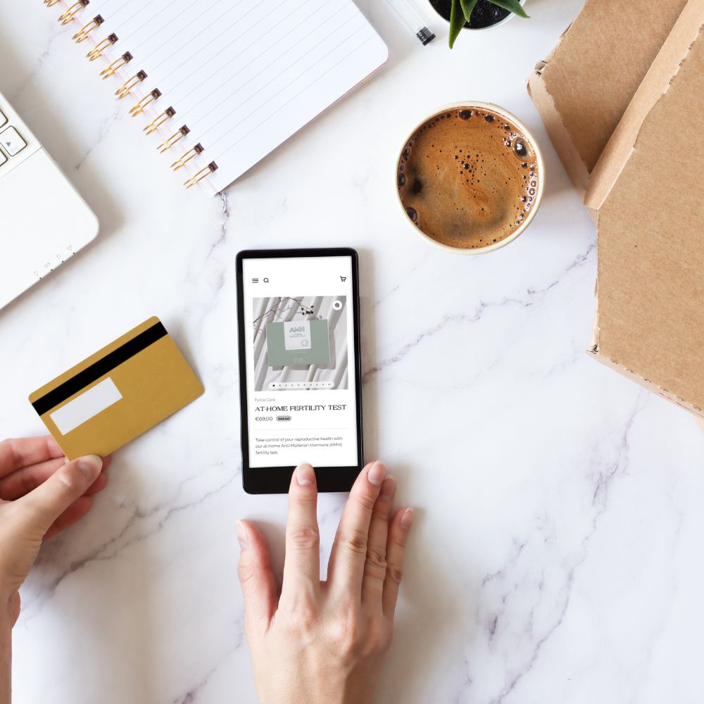 Person browsing the Fyre Care online store on a smartphone to purchase an Anti-Müllerian Hormone (AMH) fertility test with a credit card next to the phone, representing the first step of buying an at-home egg freezing fertility test.